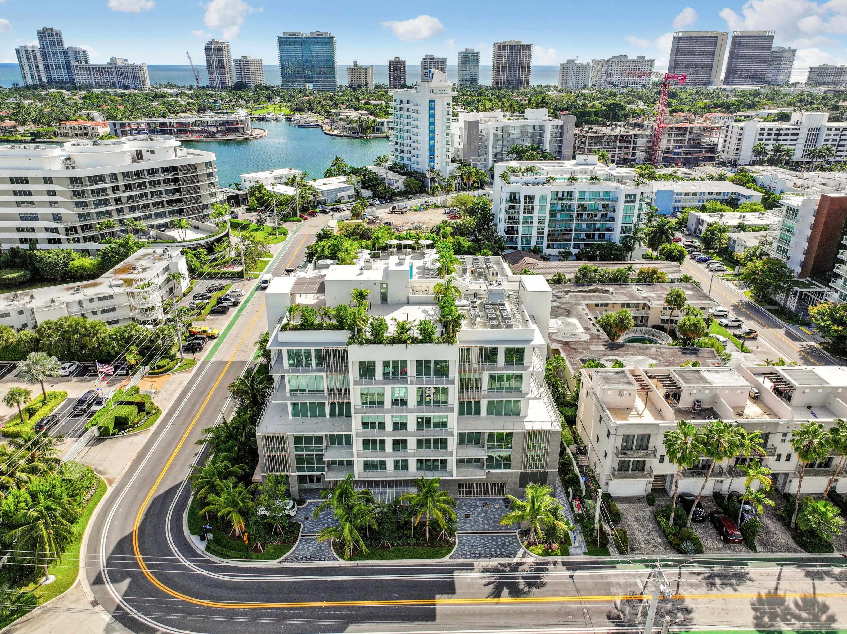 A modern building with a green roof is surrounded by other buildings and palm trees.